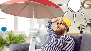 Ein Mann sitzt mit Ventilator auf der Couch, er trägt einen Hut, eine Sonnenbrille. Im Hintergrund ist ein Sonnenschirm 