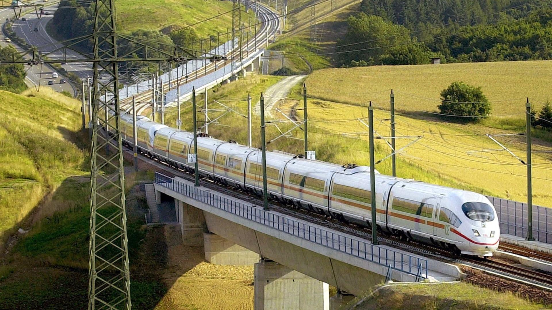 Der ICE 3 fährt auf einer Brücke durch eine Landschaft mit gelben Feldern. Im Hintergrund sieht man eine Autobahn.