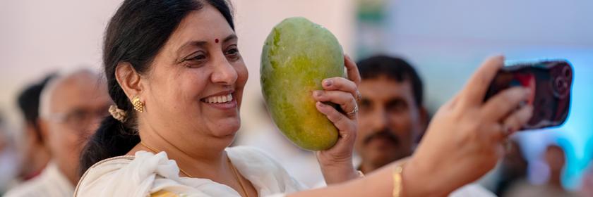 Eine Frau macht auf dem Mango-Festival ein Selfie mit einer großen Mango, die sie sich neben den Kopf hält. 