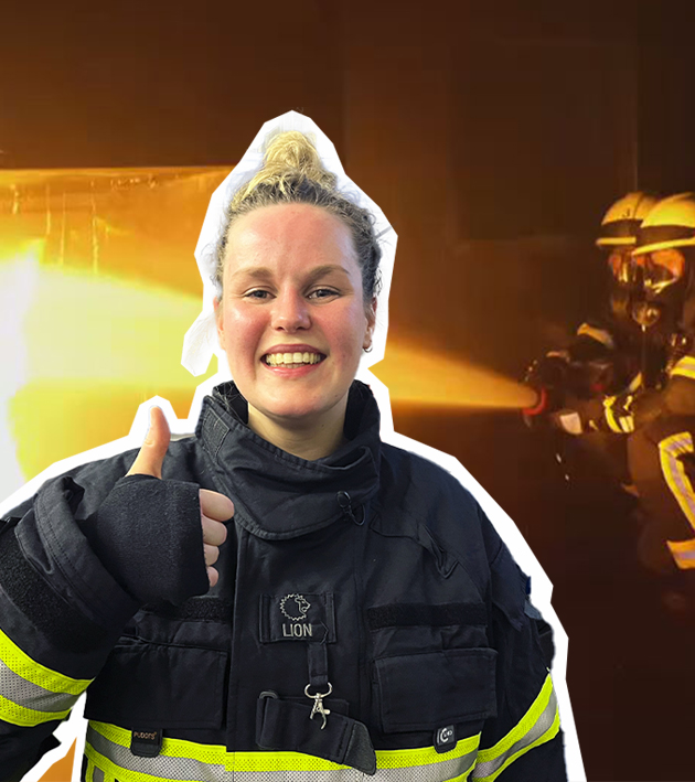 Isabelle in Feuerwehrkleidung mit Daumen hoch, im Hintergrund löschen Feuerwehrleute einen Brand.