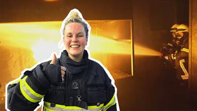 Isabelle in Feuerwehrkleidung mit Daumen hoch, im Hintergrund löschen Feuerwehrleute einen Brand.