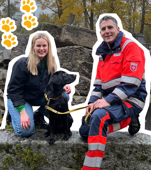 Isabelle und ein Sanitäter hocken auf einem Stein, zwischen ihnen ein schwarzer Hund, daneben Hundepfoten.