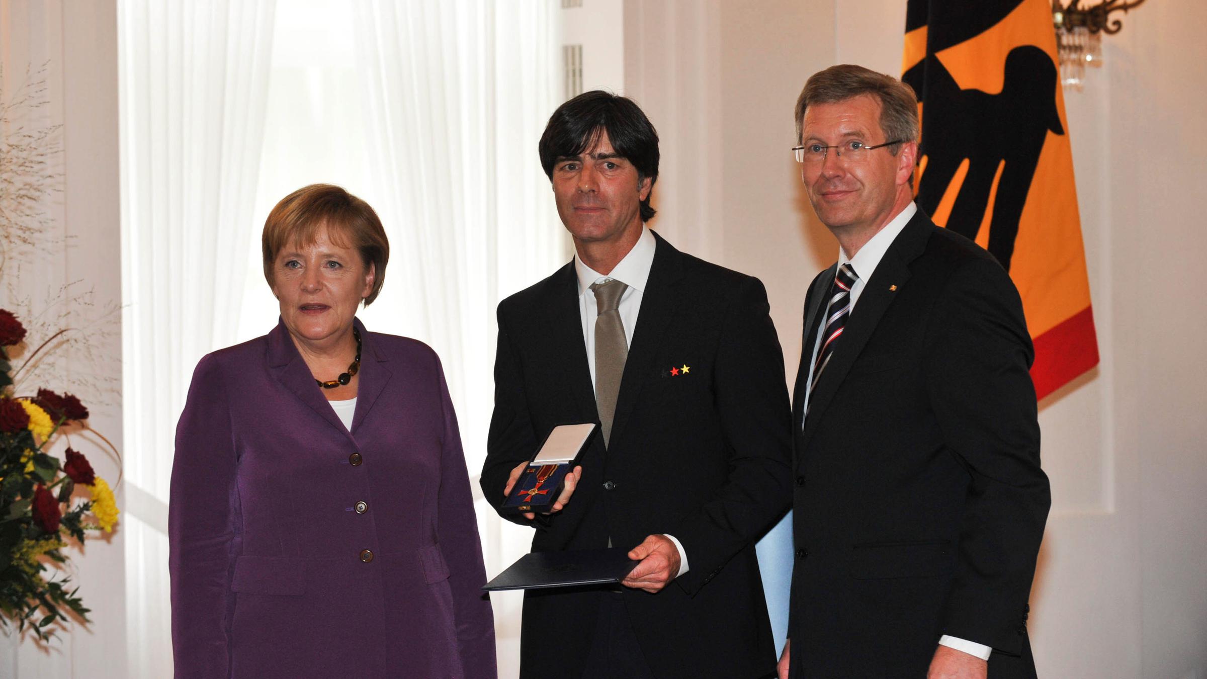 Angela Merkel, Joachim Löw und Christian Wulff stehen nebeneinander während Joachim Löw das Verdienstkreuz am Bande von Bundespräsident Christian Wulff erhält.