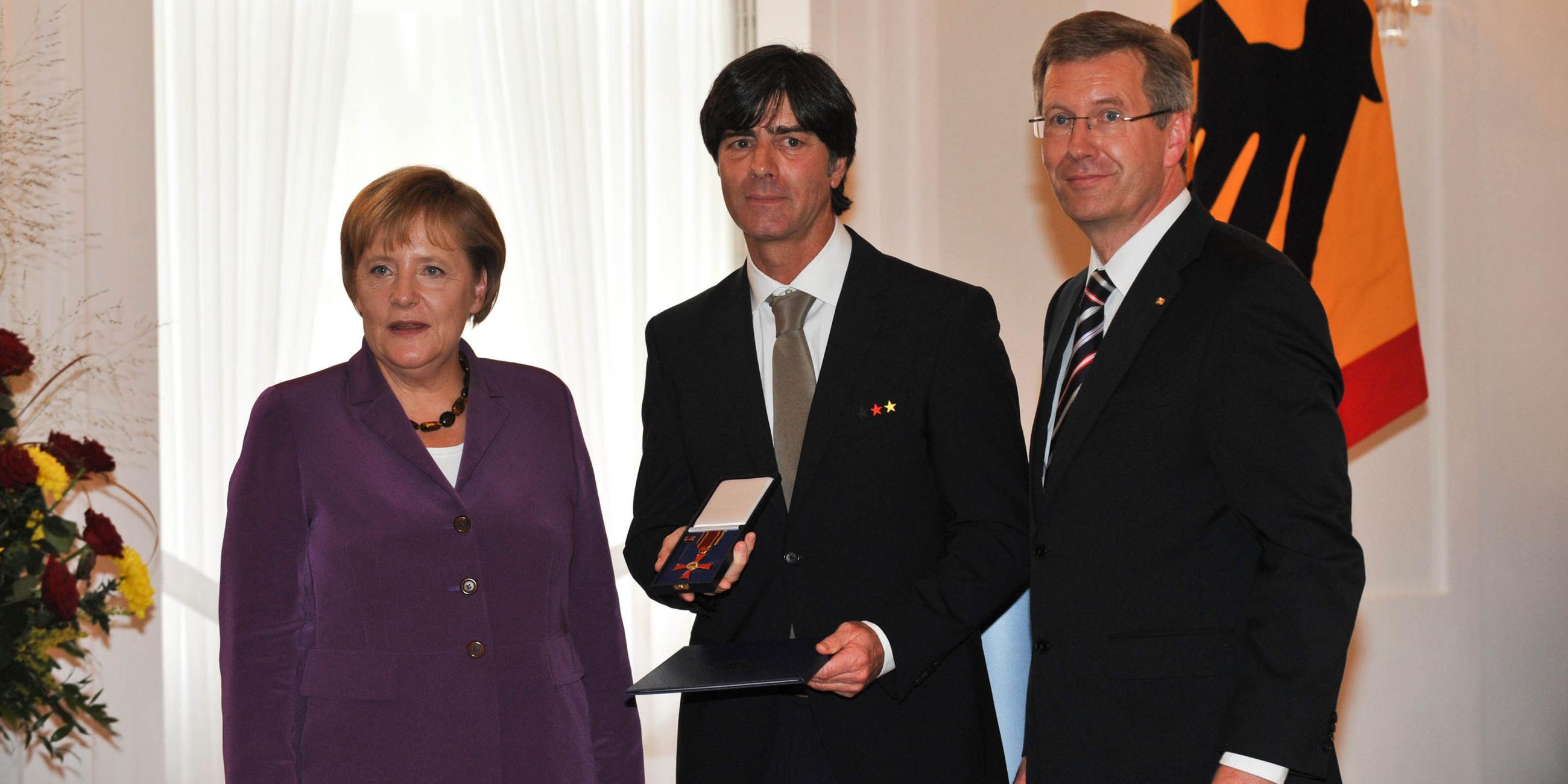 Angela Merkel, Joachim Löw und Christian Wulff stehen nebeneinander während Joachim Löw das Verdienstkreuz am Bande von Bundespräsident Christian Wulff erhält.
