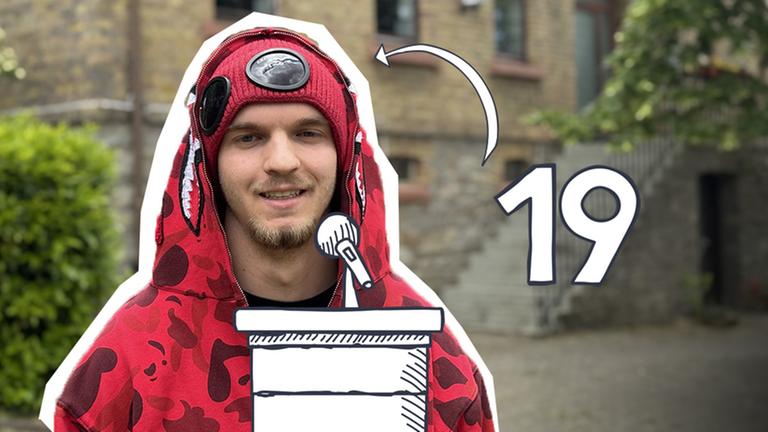 Ein junger Mann im roten Kapuzenpullover vor einem Gebäude.