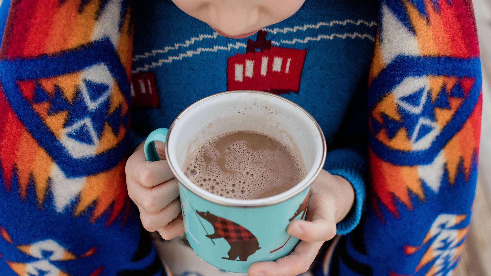 Tasse Kakao in der Hand von einem Kind, es hat eine Decke auf der Schulter 