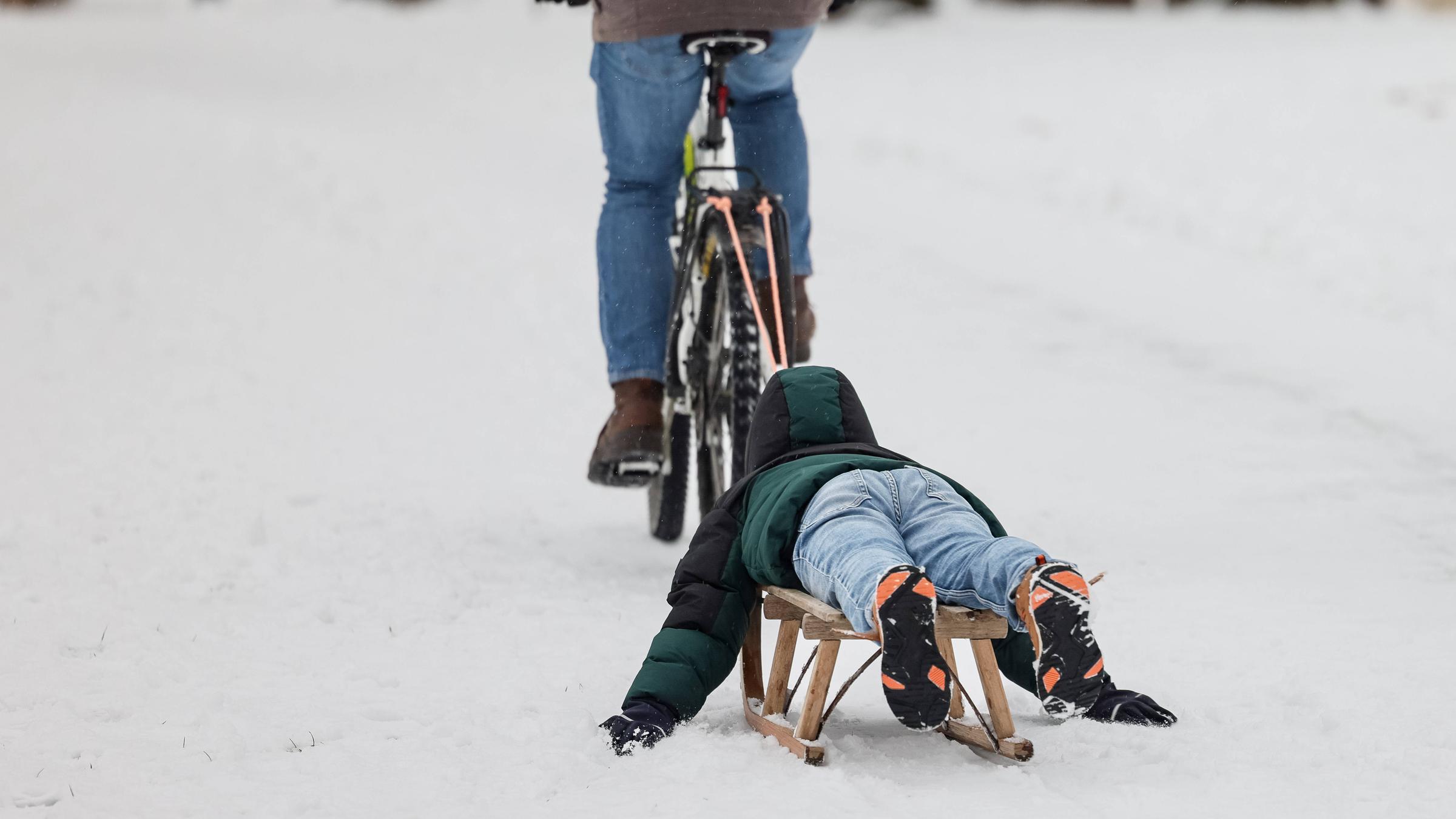 Ein Kind lässt sich am auf einem Holzschlitten von einem Fahrrad über einen verschneiten Weg am Westerberg in Osnabrück (Niedersachsen, Deutschland) ziehen. 