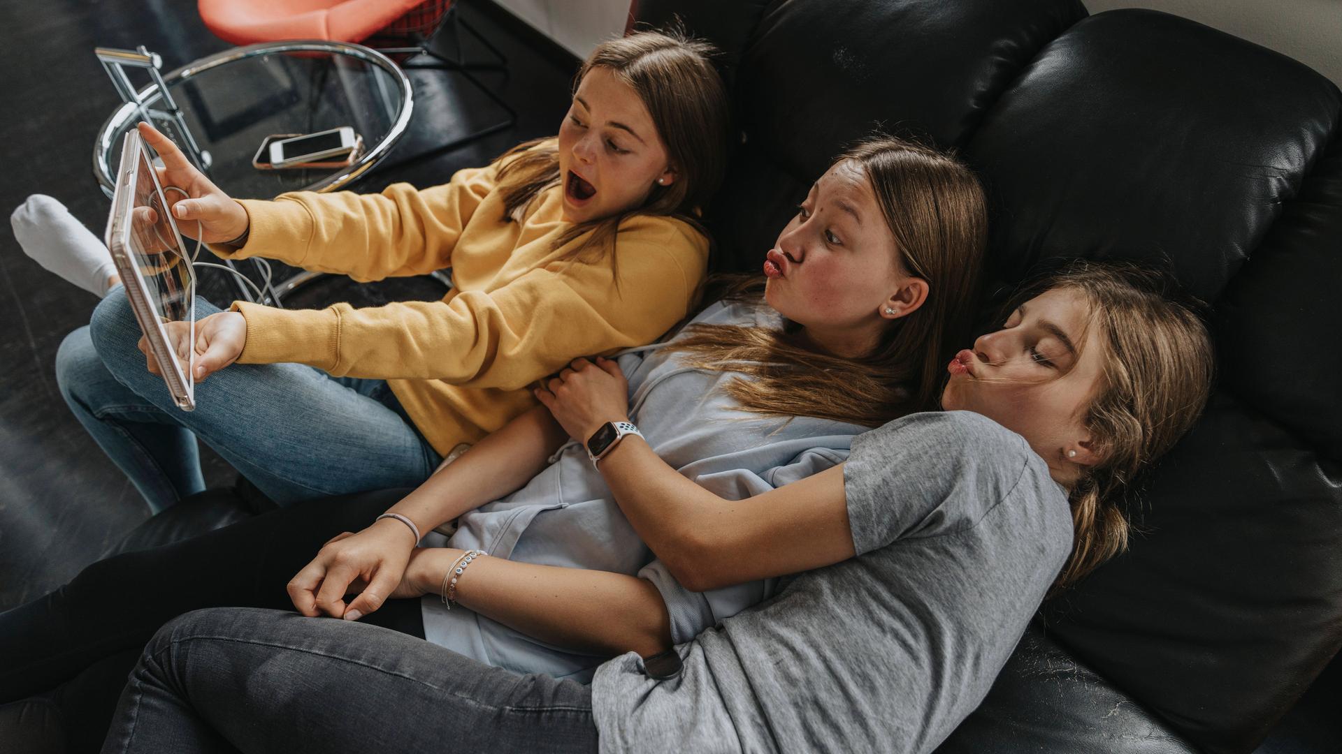 drei Mädchen sitzen auf einer Couch und ziehen Grimassen. Dabei hält eine ein Tablet in der 