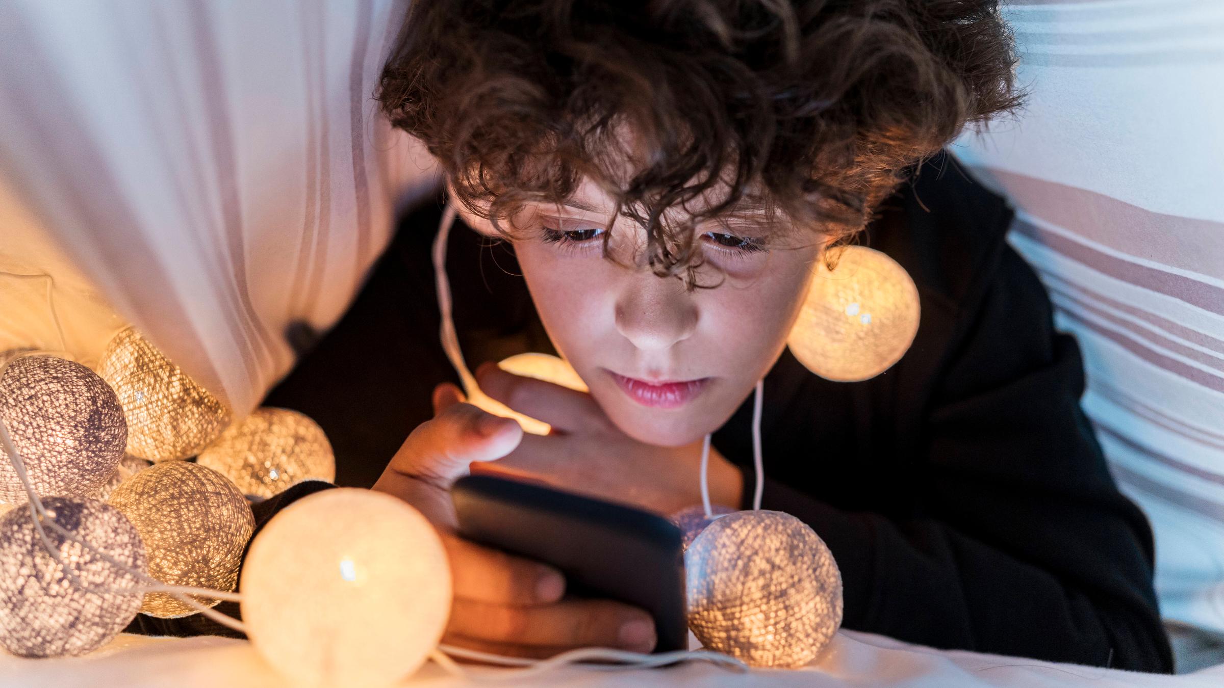Junge schaut im Bett unter der Decke ins Smartphone, trägt Lichtergirlande um den Hals