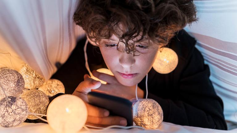 Junge schaut im Bett unter der Decke ins Smartphone, trägt Lichtergirlande um den Hals