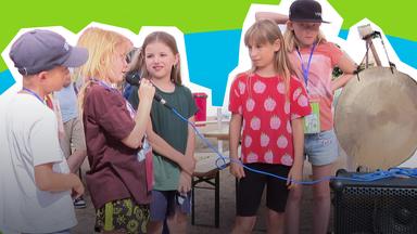 Gruppe von Kindern; ein Kind spricht in ein Mikrofon, ein anderes hält einen Gong hoch.