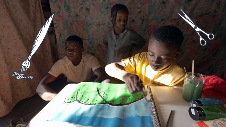 Drei Jungs an einem Tisch, einer malt ein Bild mit blauen und grünen Farben. 