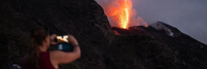 Ein Vulkan spukt Lava bei Nacht, ein Mensch fotografiert es auf Abstand.