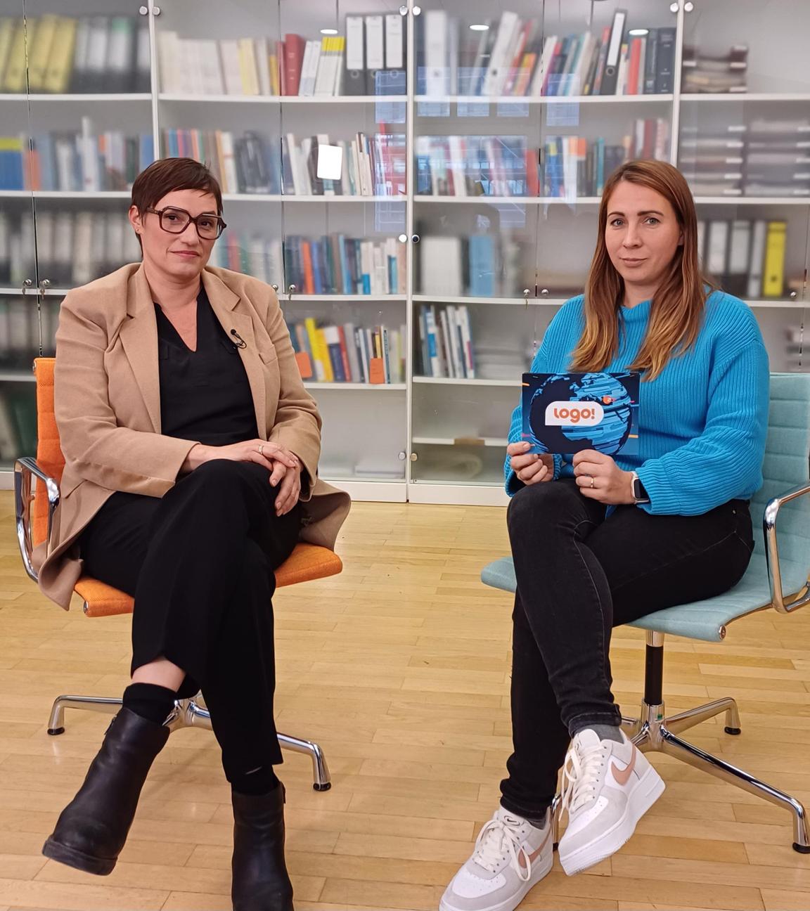 logo!-Reporterin Magdalena sitzt neben einer Expertin vor einem Bücherregal.