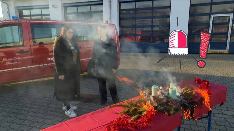 Magdalena steht neben einer Feuerwehrfrau vor einem brennenden Adventskranz