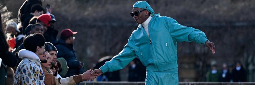 Ein männliches Model in einem türkisfarbenen Jogginganzug und Mütze und mit schwarzer Sonnenbrille. Er reicht jemandem im Publikum die Hand. Er sieht sehr cool aus. Die Menschen sind draußen in Paris vor einer Treppe.