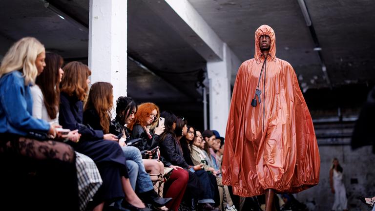 Ein Model in einem orangenen cape wir beobachtet von sitzenden Menschen links