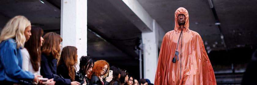 Ein Model in einem orangenen cape wir beobachtet von sitzenden Menschen links