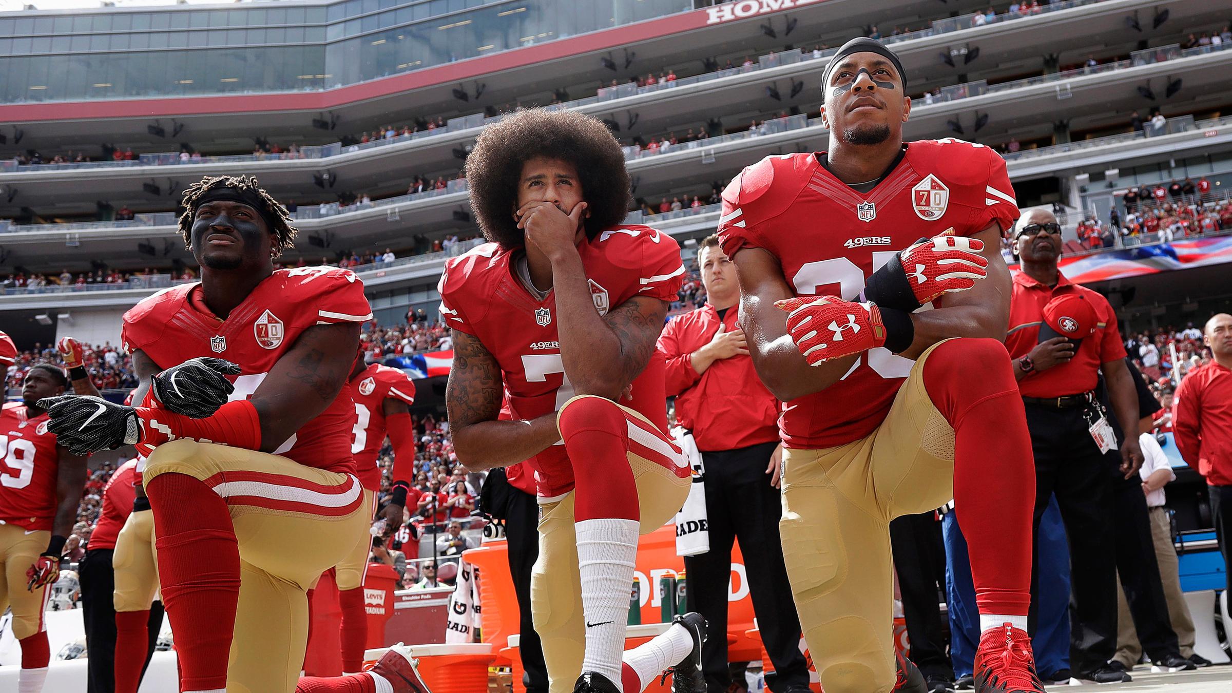 Auf dem Bild kniet der Football-Spieler Colin Kaepernick (Mitte) während der US-Nationalhymne. Mit dieser Geste protestierte er 2016 gegen Rassismus und Polizeigewalt gegen Schwarze Menschen in den USA.