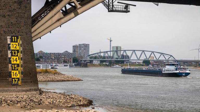Der Pfeiler eine großen Brücke ist links im Bild, er steht im Flussbett. Drumherum niedrigstehendes Wasser und in der Mitte ein Frachtschiff. Im Hintergrund ist Industrie zu sehen.