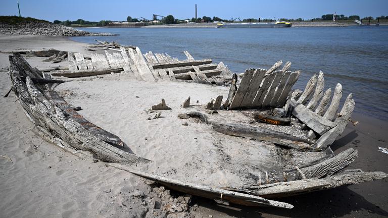 Die Holzreste eines Schiffsrumpfes, der mit Sand bedeckt ist, sind im Vordergrund zu sehen, daninter Wasser und blauer Himmel. 