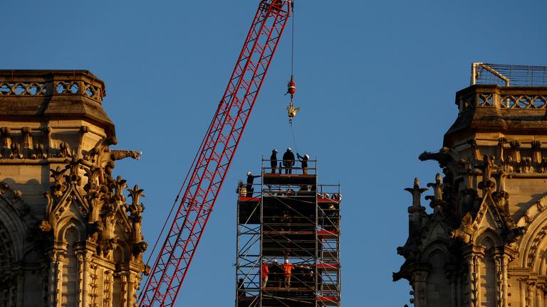 Ein Kran hebt den neuen goldenen Hahn auf die Spitze des Turms, auf der mehrere Menschen nach oben schauen.