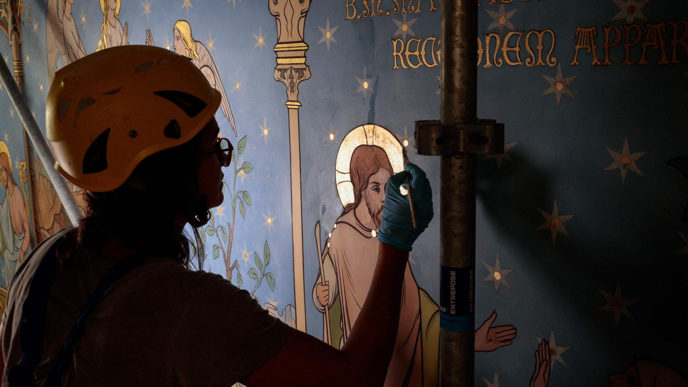 Eine Frau trägt einen Helm und restauriert mit einem Pinsel ein blaues Wandbild.