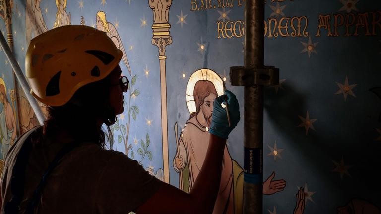 Eine Frau trägt einen Helm und restauriert mit einem Pinsel ein blaues Wandbild.