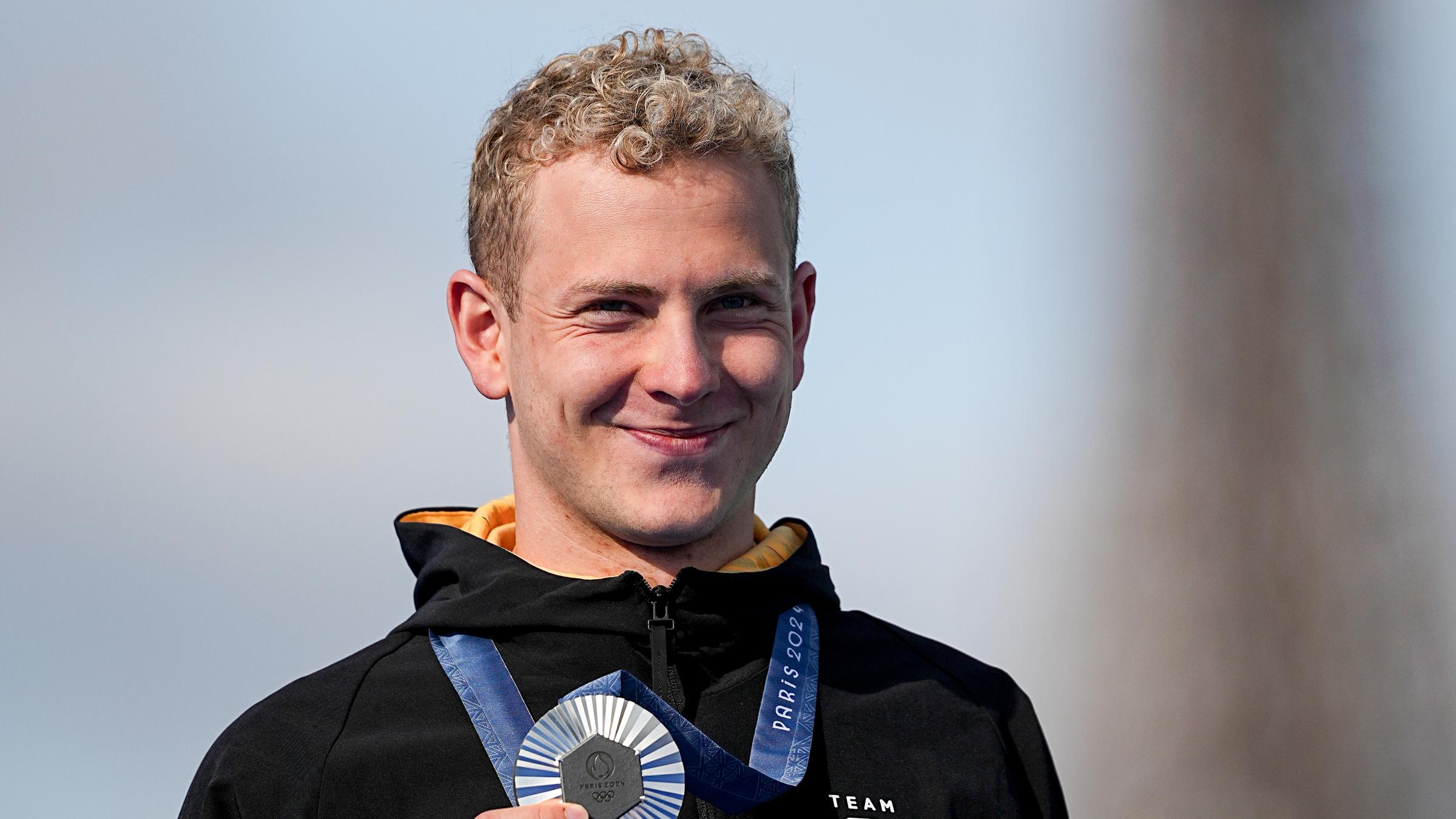 Blonder Mann lächelt in die Kamera und hält seine Silbermedaille in der Hand. 