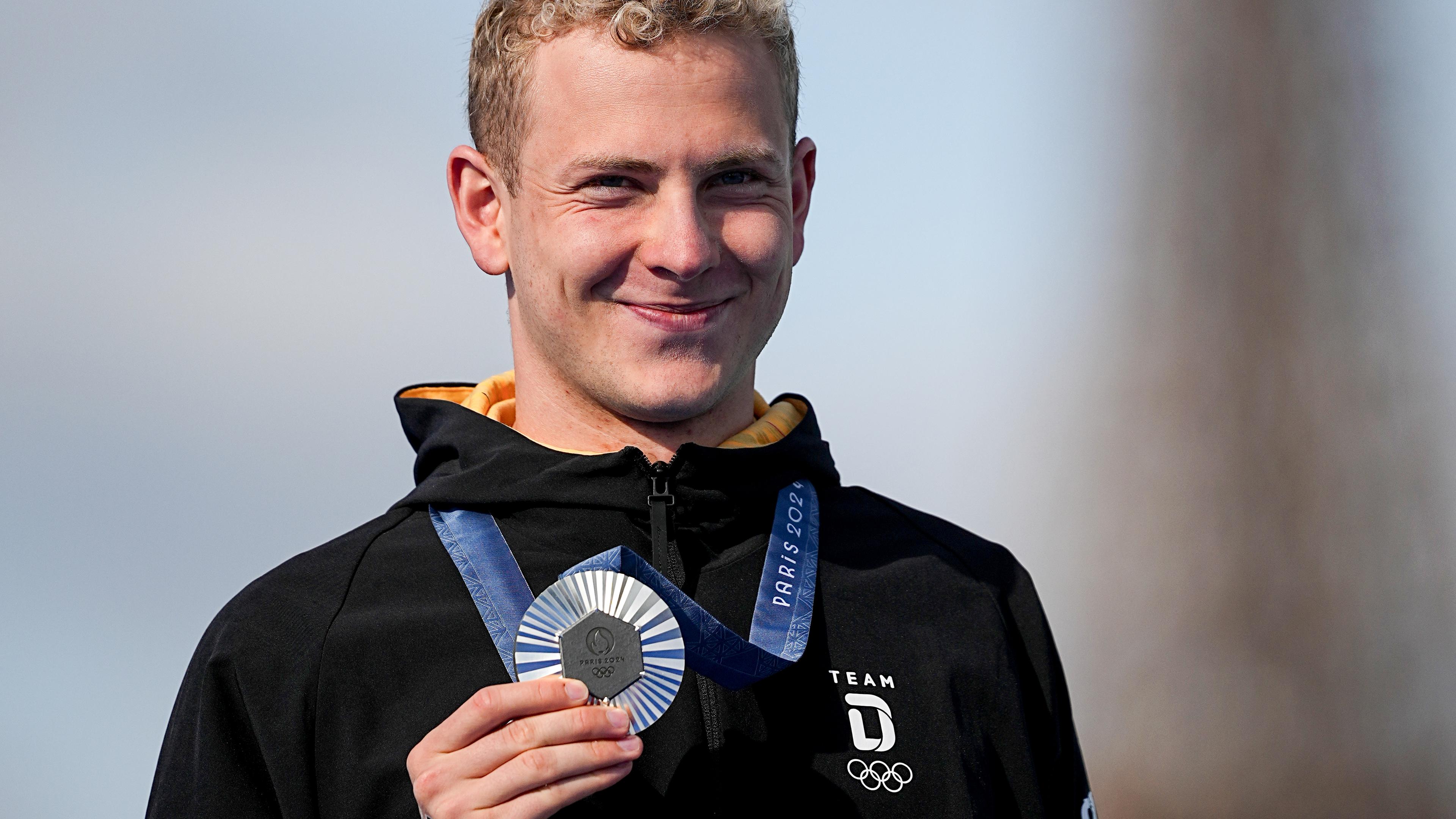 Blonder Mann lächelt in die Kamera und hält seine Silbermedaille in der Hand. 