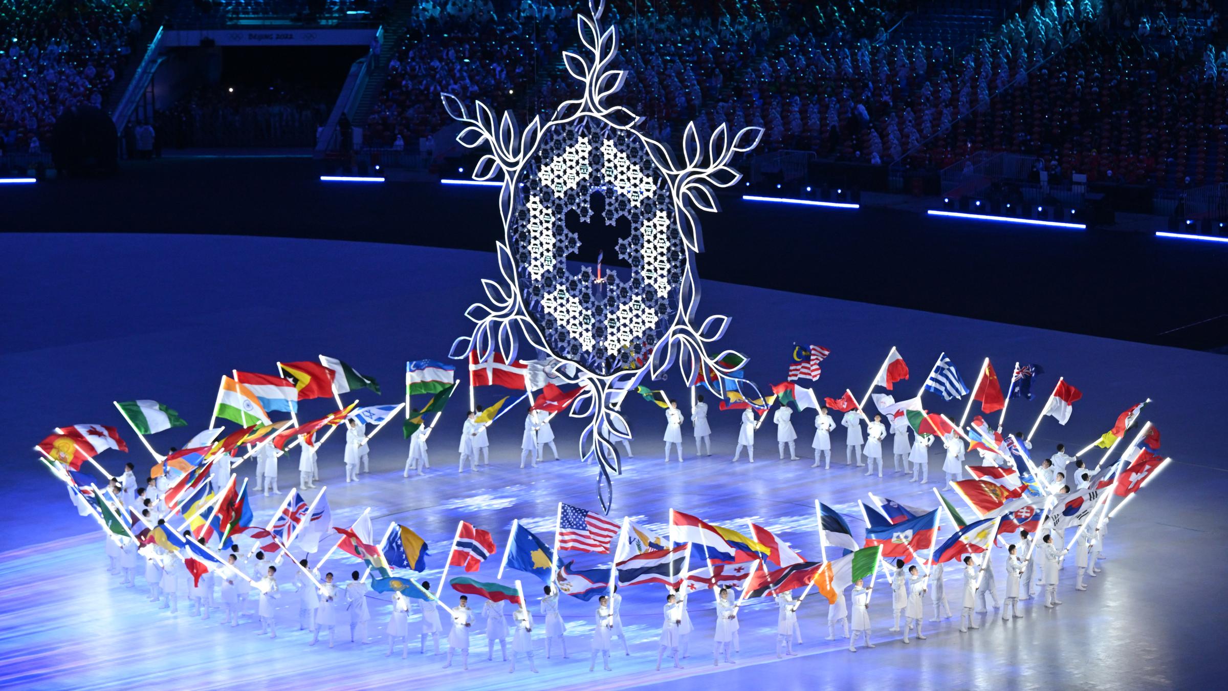 Schlussfeier der Olympischen Spiele mit Fahnenträgern, die im Kreis um eine riesige Skulptur einer stilisierten Schneeflocke herumgehen. 