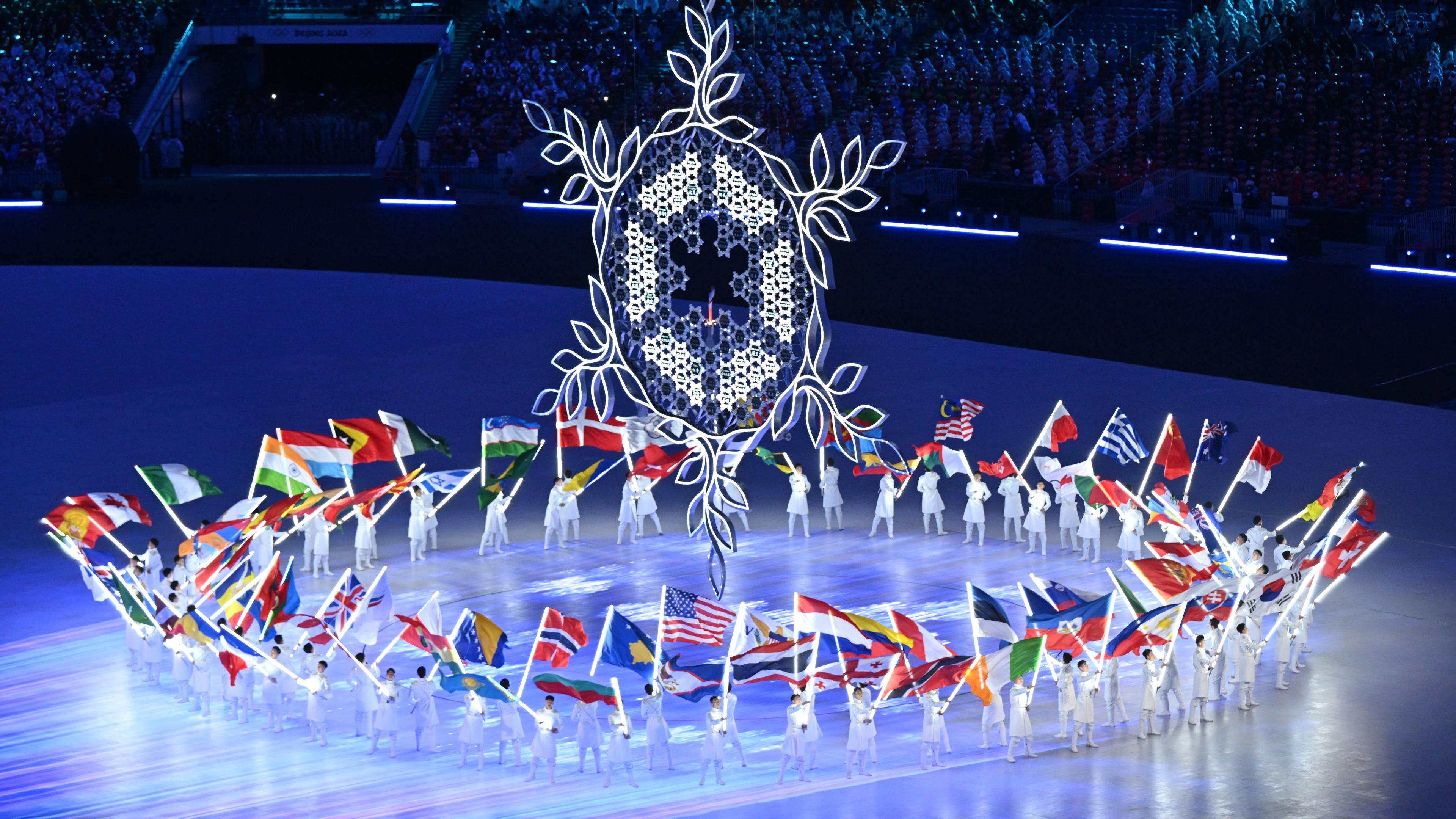 Schlussfeier der Olympischen Spiele mit Fahnenträgern, die im Kreis um eine riesige Skulptur einer stilisierten Schneeflocke herumgehen. 