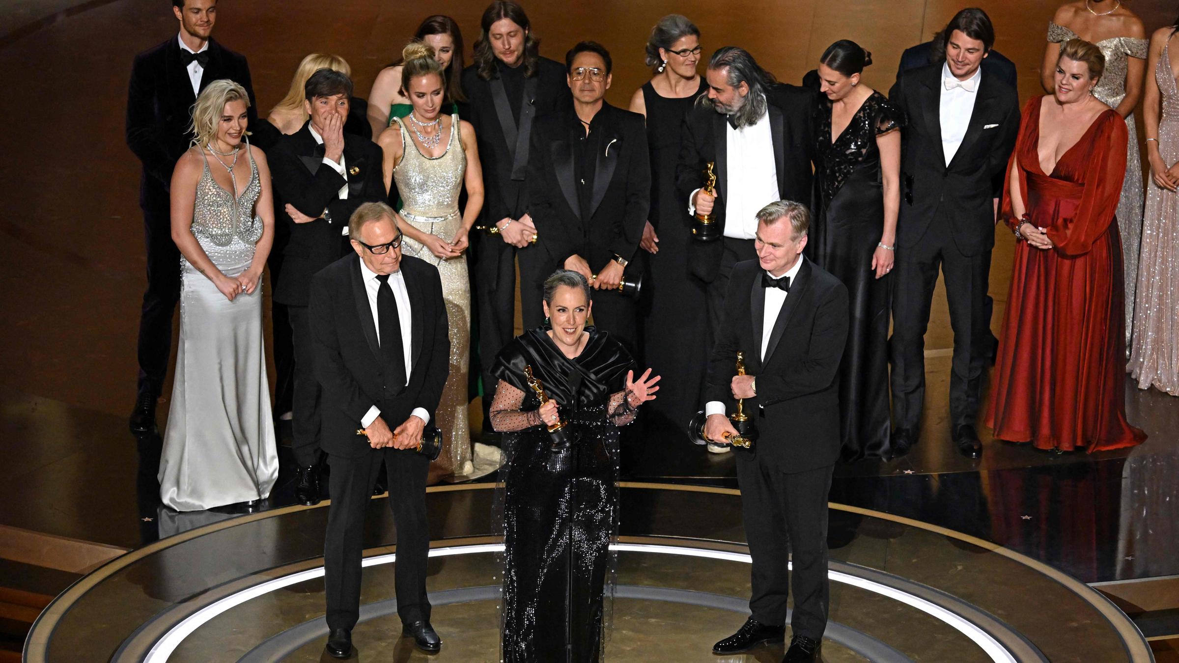 Der Regisseur Christopher Nolan, die Filmproduzentin Emma Thomas und der Filmproduzent Charles Roven bei ihrer Dankesrede für den besten Film. Im Hintergrund sind weitere Menschen, die bei dem Film beteiligt waren, auf der Bühne zu sehen.