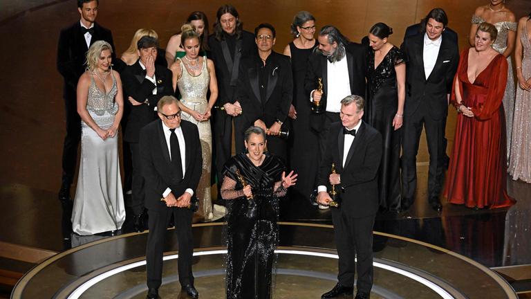 Der Regisseur Christopher Nolan, die Filmproduzentin Emma Thomas und der Filmproduzent Charles Roven bei ihrer Dankesrede für den besten Film. Im Hintergrund sind weitere Menschen, die bei dem Film beteiligt waren, auf der Bühne zu sehen.