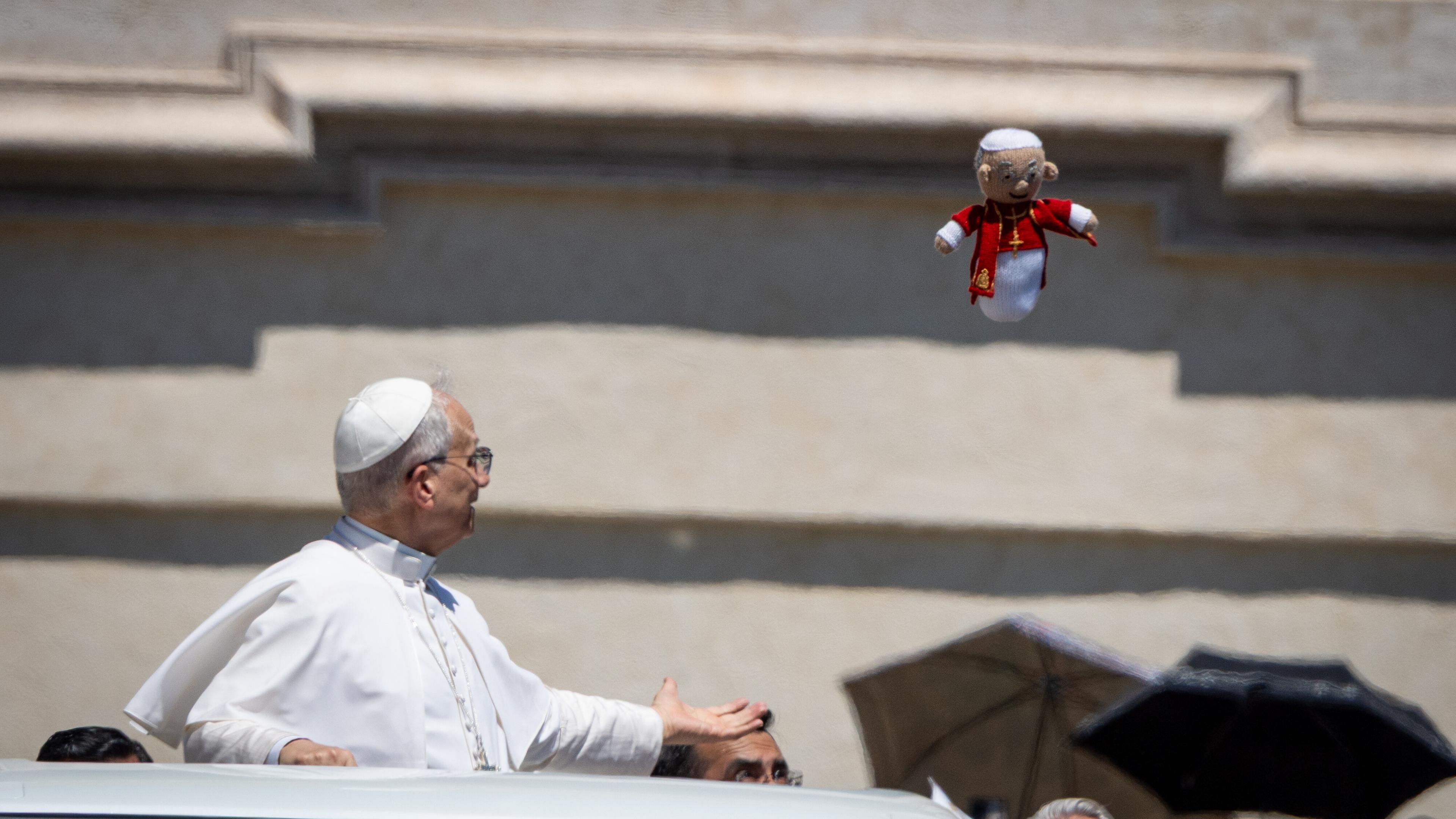 Papst Leo XIV. steht in einem offenen Fahrzeug und schaut nach oben. In der Luft fliegt eine kleine Stoffpuppe, die aussieht wie ein Mini-Papst mit rotem Mantel.