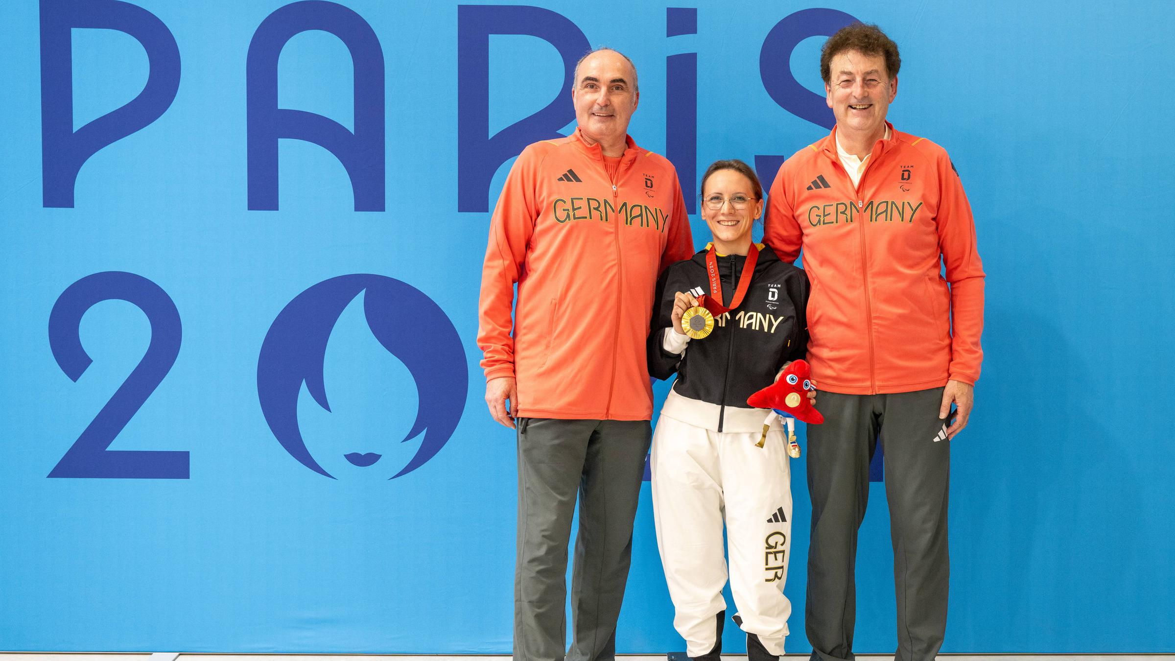 Natascha Hiltrop steht zwischen zwischen ihren beiden Coaches mit der Goldmedaille in der Hand