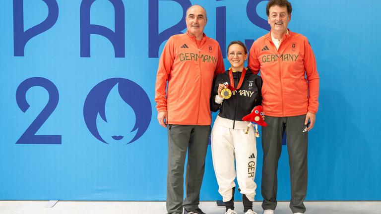Natascha Hiltrop steht zwischen zwischen ihren beiden Coaches mit der Goldmedaille in der Hand