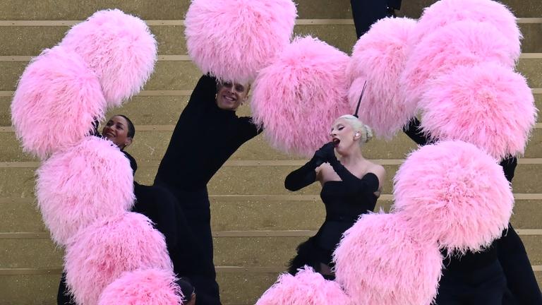 Lady Gaga singt und um sie herum wird ein Herz aus pinkfarbenen Puscheln geformt. 