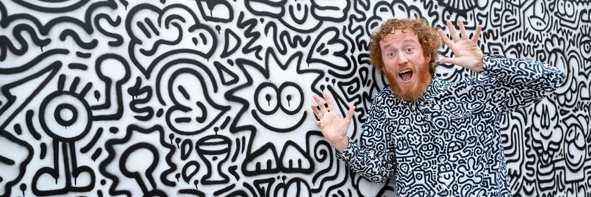 Ein Künstler steht neben seinem Kunstwerk. Seine Kleidung hat das gleiche Muster wie das Kunstwerk. 