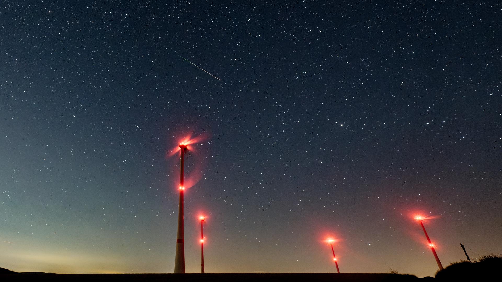 Blick in den Nachthimmel. Man sieht Windräder mit rotleuchtenden Lampen, die Milchstraße, die wolkig aussieht und einen weißen Strich, das ist eine Sternschnuppe
