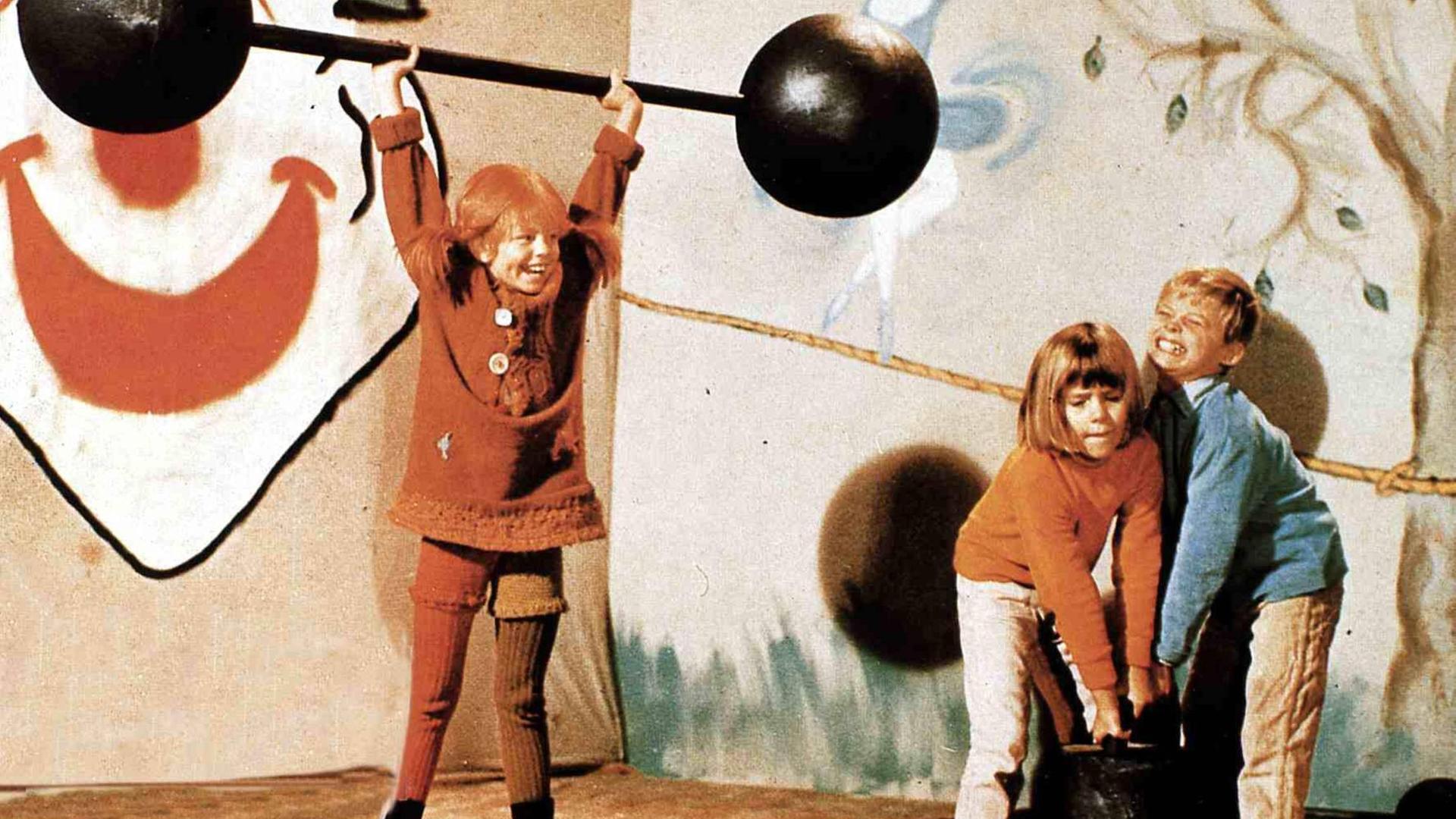 Pippi Langstrumpf hebt ein großes Gewicht über ihren Kopf und Annika und Tommy versuchen, ein Gewicht auf dem Boden hochzuheben, aber schaffen es nicht.