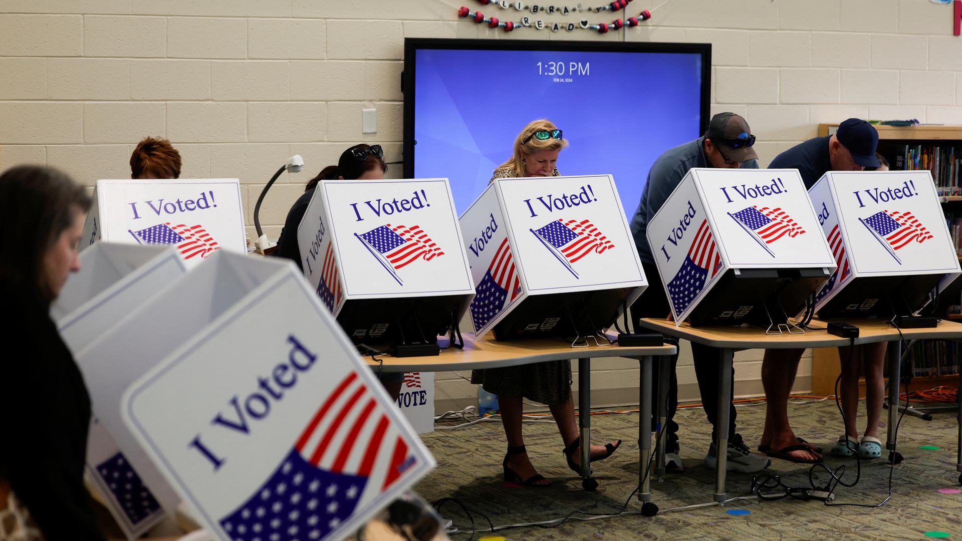 Mehrer Menschen stehen nebeneinander und stimmen in kleinen Wahlkabinen ab. Auf den Wahlkabinen steht "I voted" und die Flagge der USA ist abgebildet.
