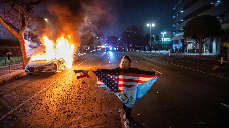 Eine Demonstrantin mit mehreren Flaggen läuft vor einem brennenden Auto