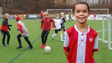 Auf einem Rasenfußballplatz. Im Vordergrund rechts steht der 10-jährige Tyler, in der Trainingskleidung des 1. FC Kaiserslautern. Hintergrund trainieren andere Kinder.