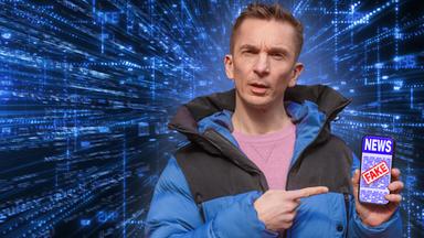 Eric trägt eine blaue Daunenjacke und darunter einen rosafarbenen Pulli. Er hält ein Smartphone in seiner linken Hand. Auf dem Display sieht man das Wort "News" und darüber ist ein roter Fake-Stempel.