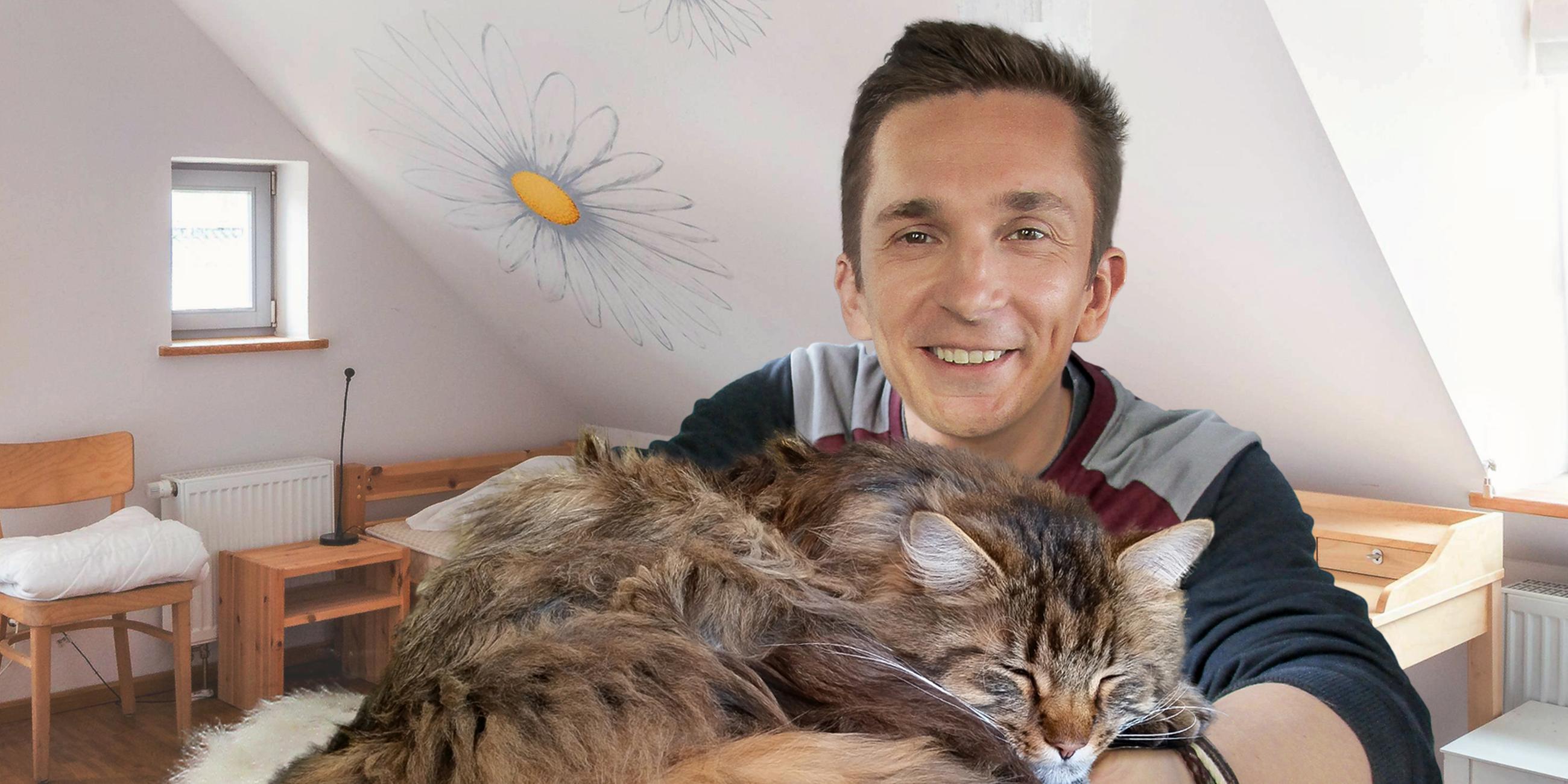 Moderator Eric Mayer lächelt in die Kamera. In seinen ineinander verschränkten Armen liegt eine Langhaar-Katze mit geschlossenen Augen und einem zufriedenen Gesichtsausdruck. Im Hintergrund ist ein Zimmer mit weißen Wänden und einem hölzernen Bett, einem Nachttisch, einem Stuhl und einem Schreibtisch zu sehen.