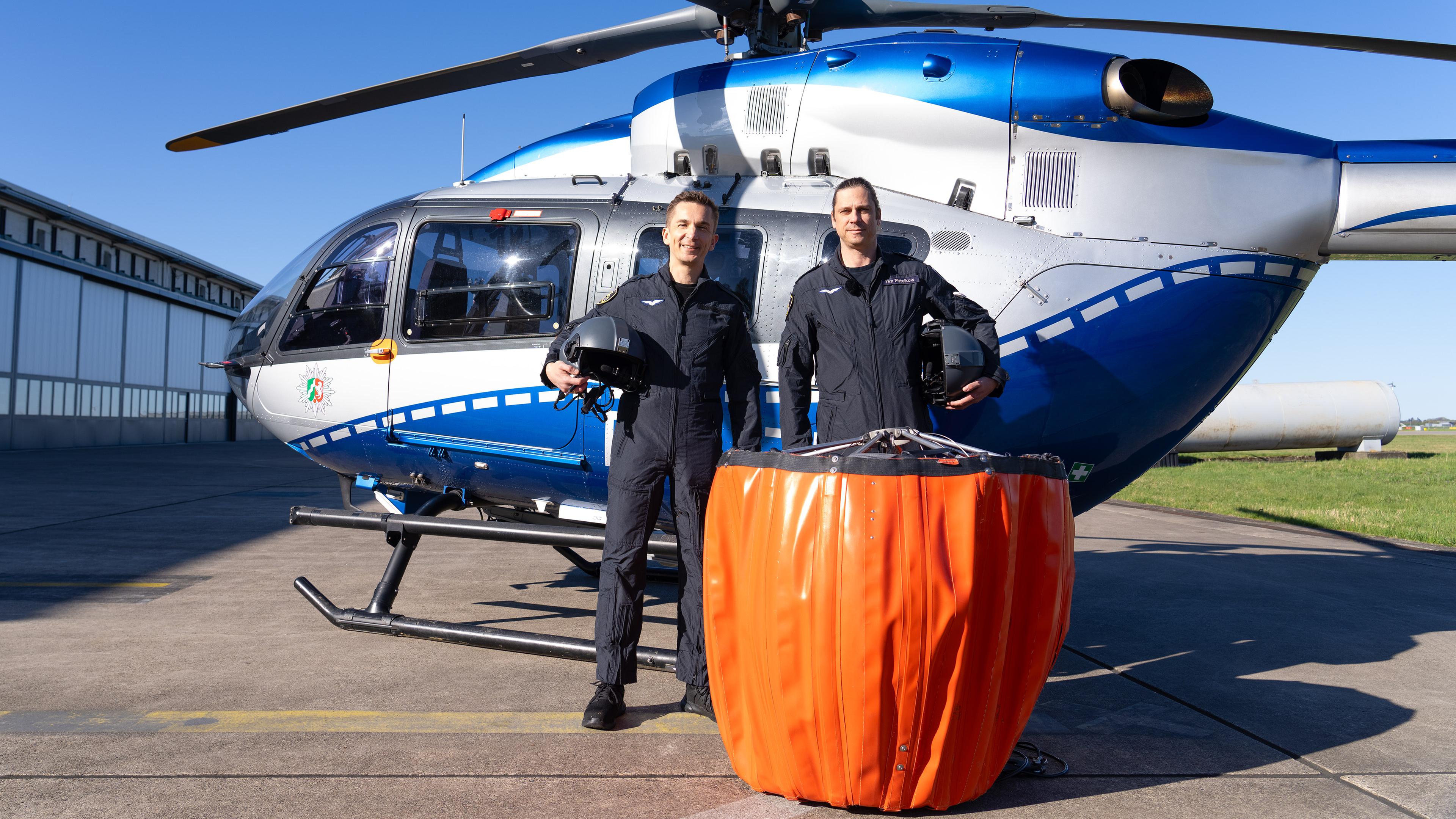 Eric Mayer steht neben Pilot Tim vor einem Hubschrauber der Polizeifliegerstaffel Nordrhein-Westfalen. Vor ihnen steht ein Außenlastbehälter, der beim Löscheinsatz unter dem Hubschrauber angehängt wird, um Wasser aus Flüssen oder Seen zu entnehmen und zu den Bränden zu transportieren.