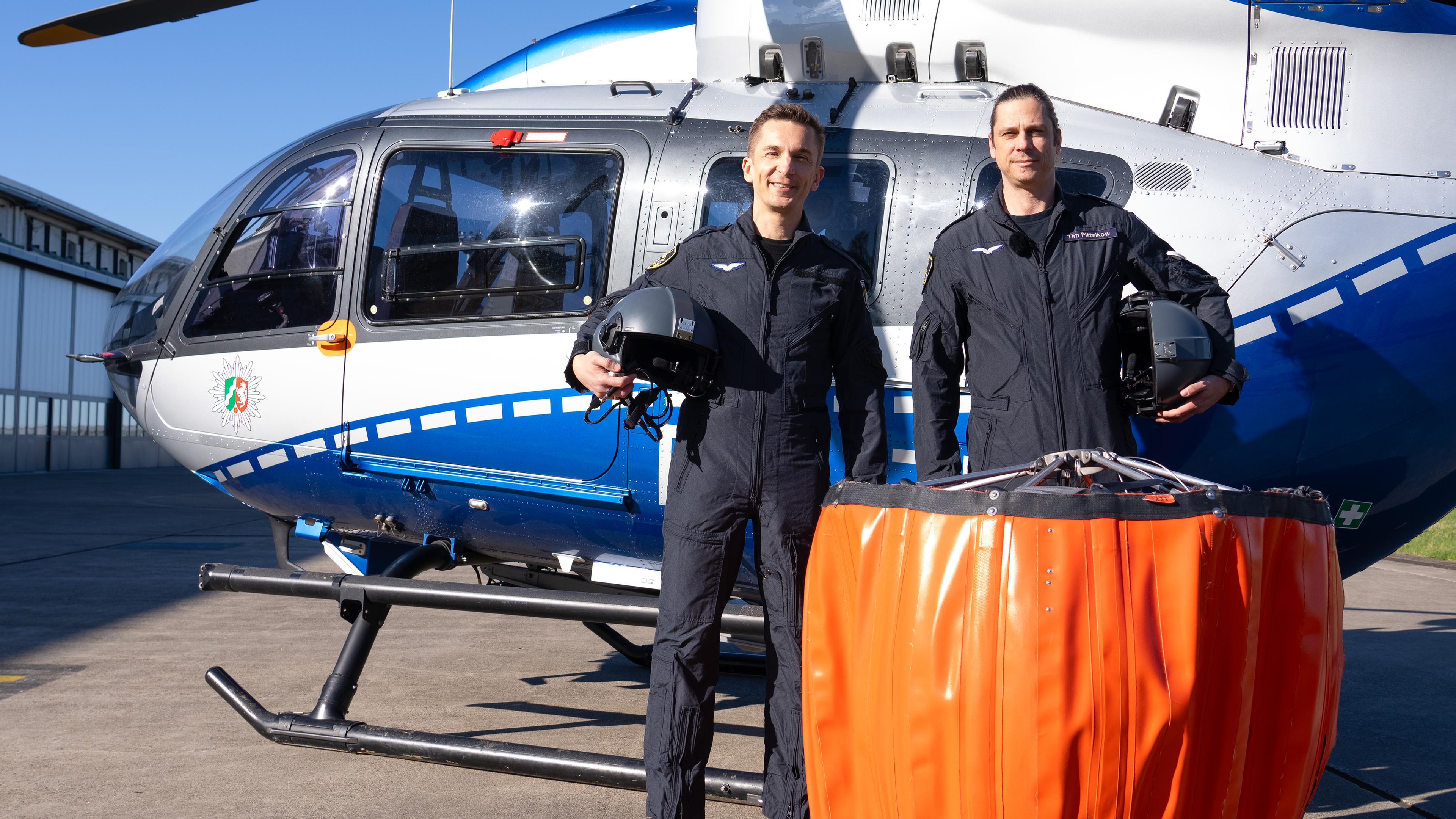 Eric Mayer steht neben Pilot Tim vor einem Hubschrauber der Polizeifliegerstaffel Nordrhein-Westfalen. Vor ihnen steht ein Außenlastbehälter, der beim Löscheinsatz unter dem Hubschrauber angehängt wird, um Wasser aus Flüssen oder Seen zu entnehmen und zu den Bränden zu transportieren.