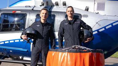Eric Mayer steht neben Pilot Tim vor einem Hubschrauber der Polizeifliegerstaffel Nordrhein-Westfalen. Vor ihnen steht ein Außenlastbehälter, der beim Löscheinsatz unter dem Hubschrauber angehängt wird, um Wasser aus Flüssen oder Seen zu entnehmen und zu den Bränden zu transportieren.