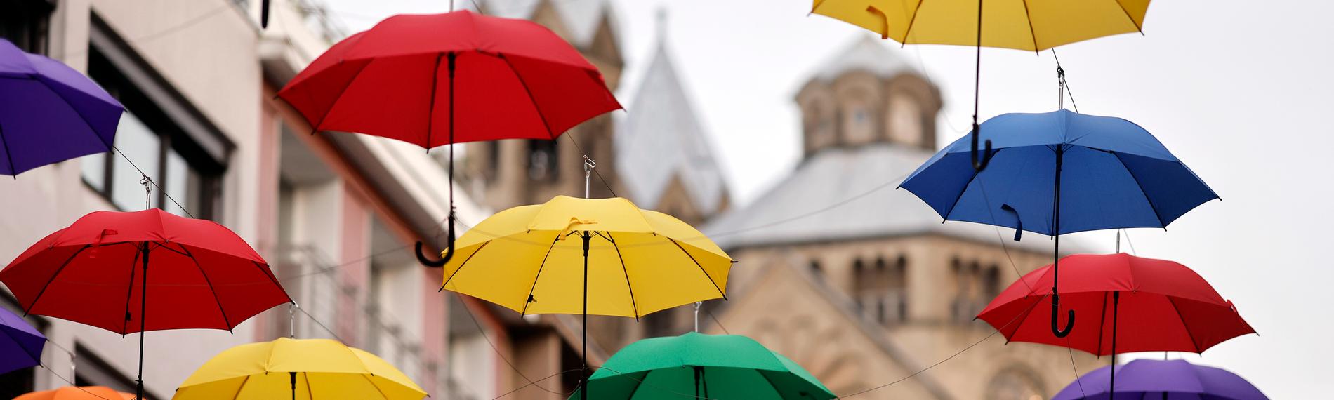Aufgespannte, bunte Regenschirme hängen zwischen zwei Gebäuden an Schnüren, es sieht aus als würden sie schweben.